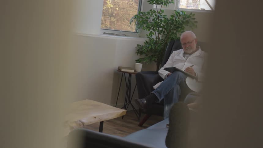 A doctor sits on a chair in an office writing on a pad of paper while a woman lays on a sofa