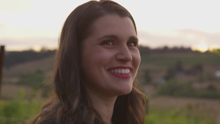 A woman is smiling as she stands outside in a vineyard at sunset