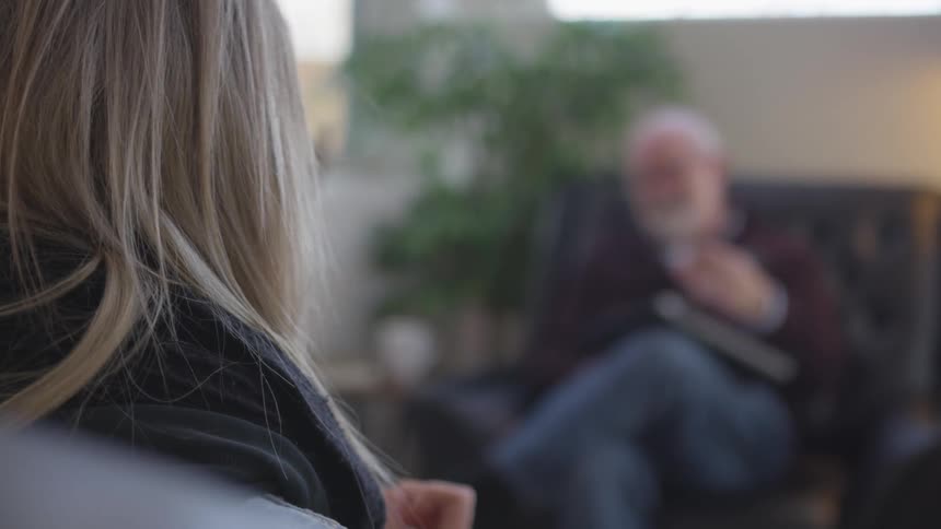 A man sits in a chair holding a pad and pen across from a man and a woman in an office
