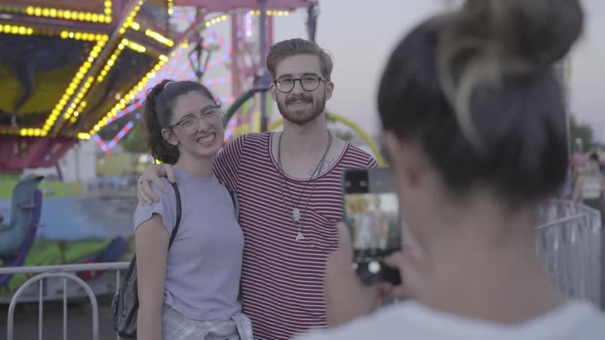 A woman is taking a photo of a couple at a fair at dusk