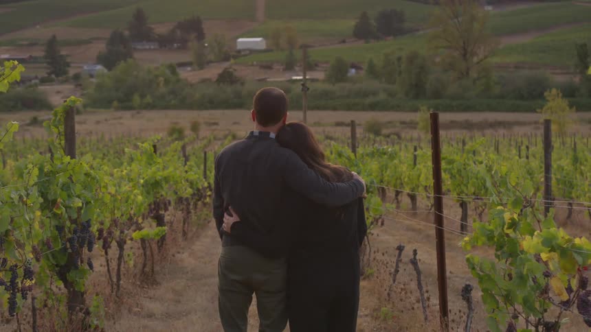 A man and woman stand an embrace as they look over a large piece of farm land