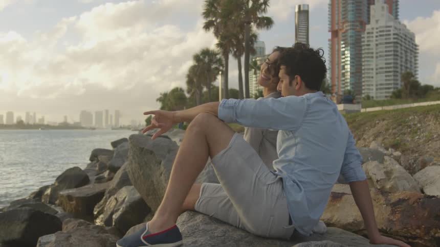 A man and a woman smile as they sit on rocks by a large body of water with clouds and buildings in the background