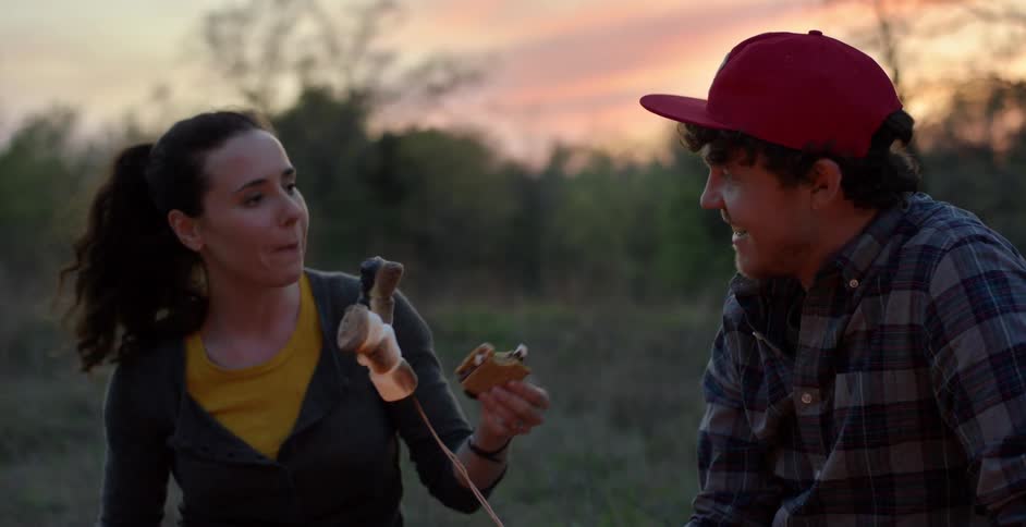 A woman feeding a man as he roasts marshmallows on a fire at dusk
