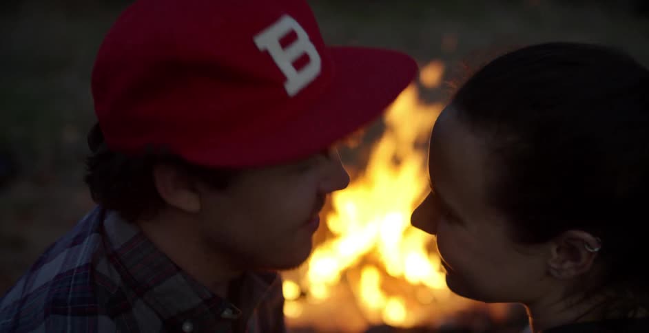 Man and woman talk, and he kisses her forehead by a campfire