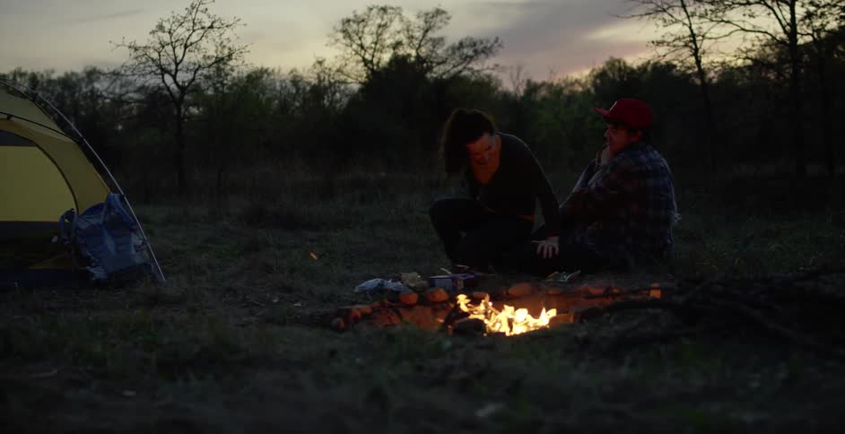 A woman brings a blanket over to a campfire at dusk to join her friend