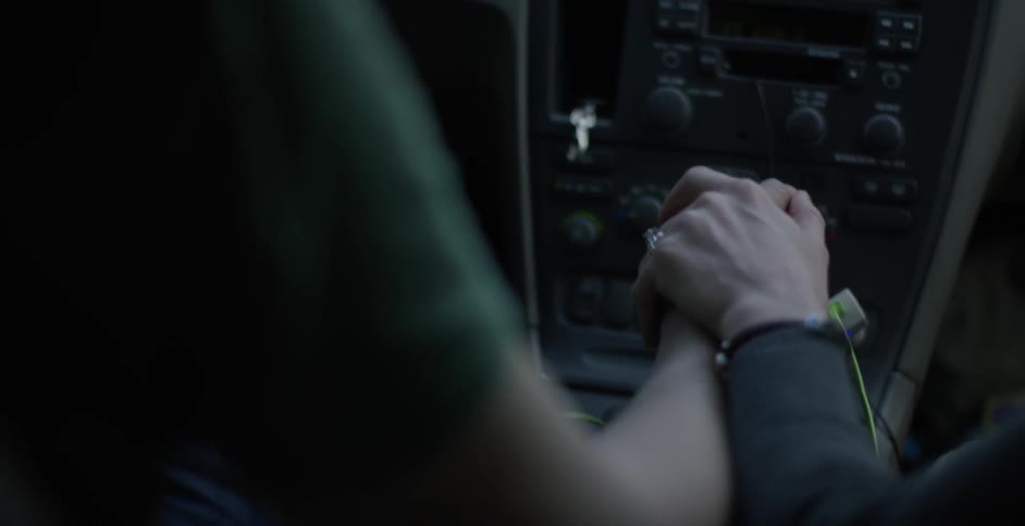 While in a car, a passenger places their hand on top of the driver's hand, which is resting on the stick shift