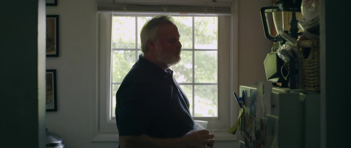 A man stands in a kitchen at day time drinking a glass of water and then walks away