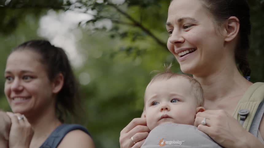 Two women holding babies in baby carriers smile as they stand outside in front of trees