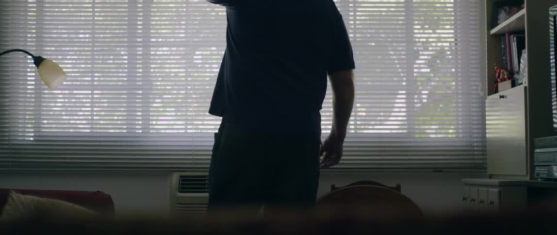 A man walks towards a window and looks outside with his hands on his hips