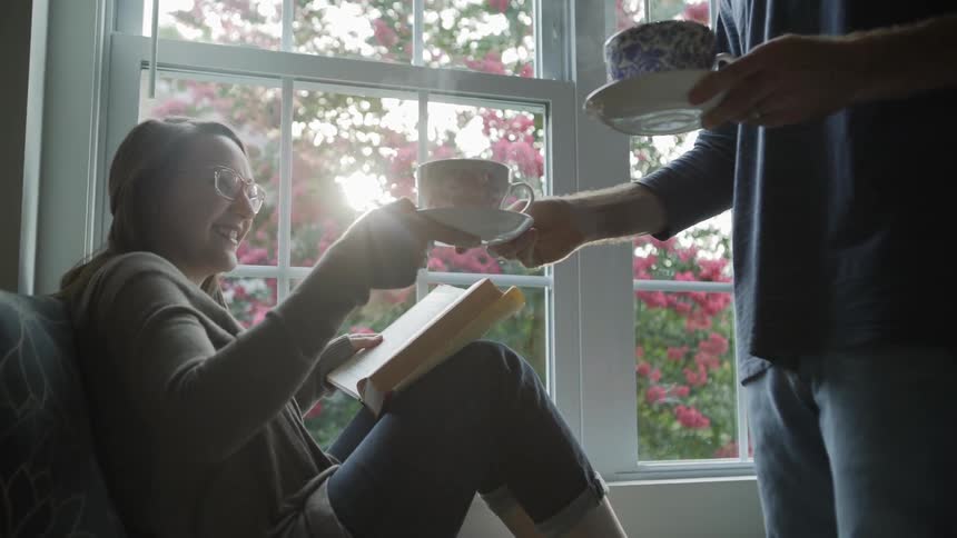 Man brings a cup of hot liquid to a woman reading as she sits on the sofa