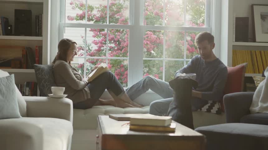 Man and woman sit on a couch next to a window and a dog stands on the ground by the man