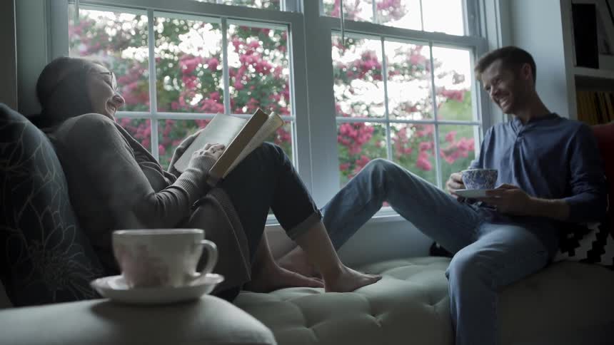 A man and a woman sit in a window seat smiling as they read and drink from tea cups