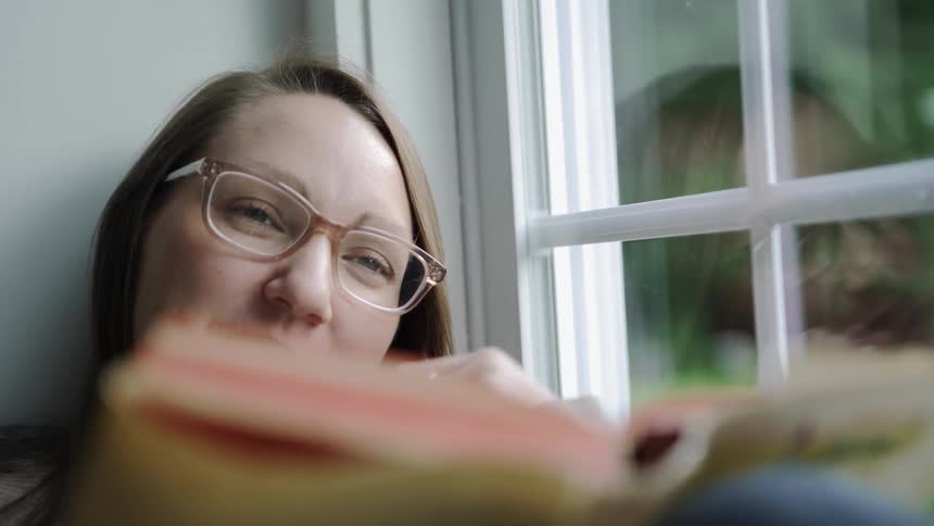 A woman sitting next to a window smiles as she holds a book