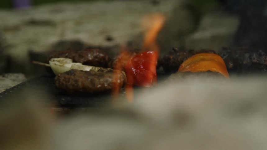 A person flips burgers and veggies cooking on a grill with flames