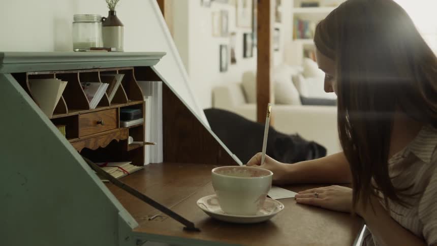 A woman sits at a desk writing and her dog walks into the room