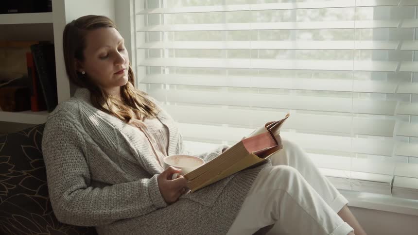 Woman sits on a couch next to a window reading a book