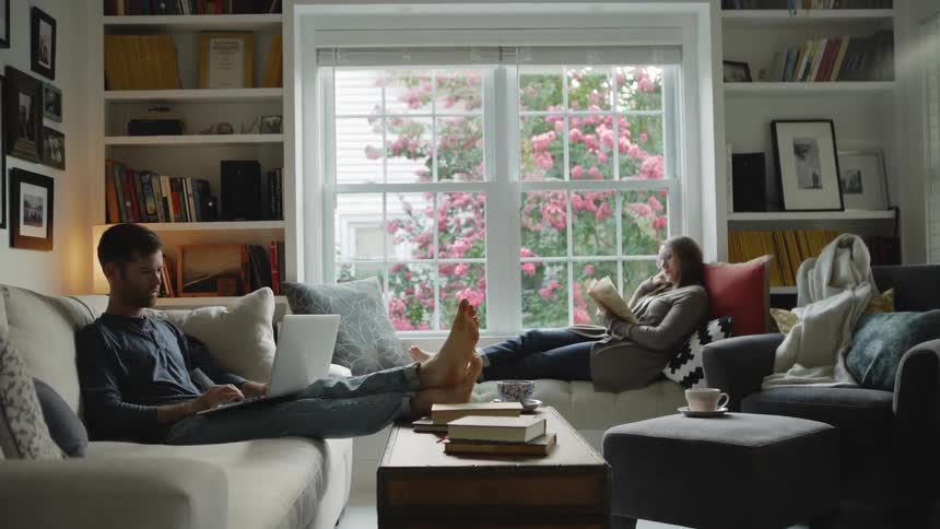 A man and woman sit in a living room reading and working on a laptop