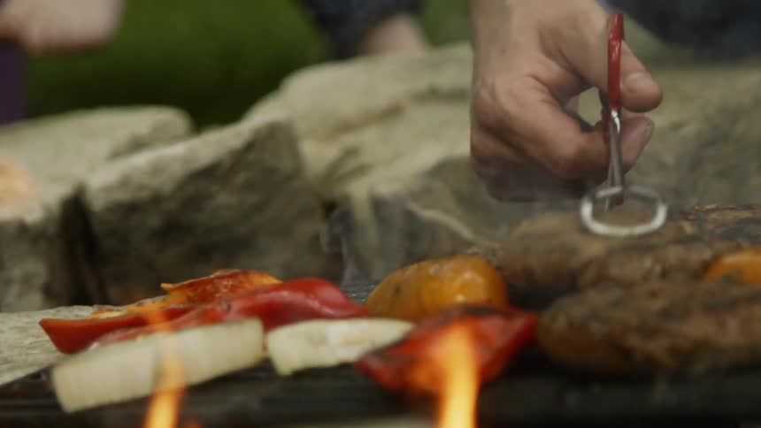 A man uses tongs to move meat and veggies around on a grill outside