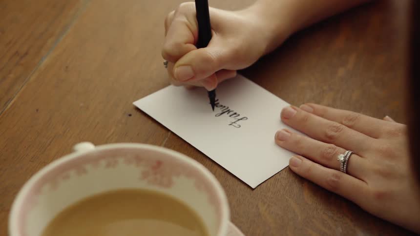 A woman sits at a desk with a cup of coffee and writes on an envelope