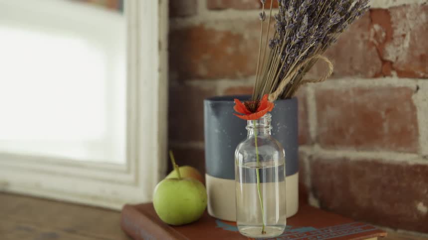 A book sits on a shelf by a window with a piece of fruit a glass bottle and a can with flowers on top of it