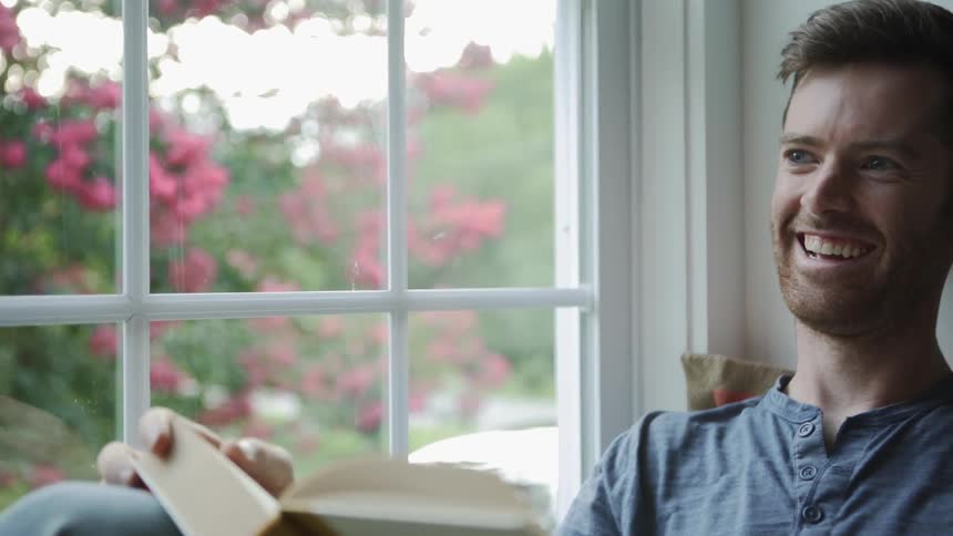 A man sits in a window seat holding a book and laughing