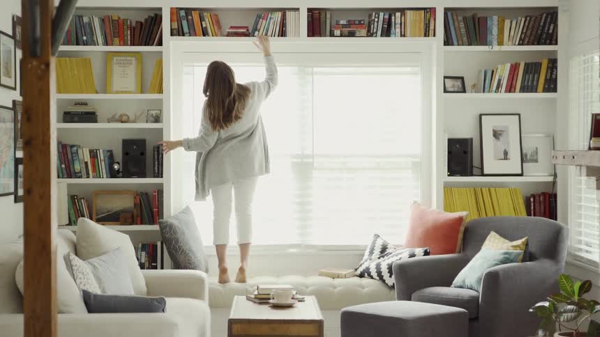 Woman stands on a sofa and reaches up into the bookshelf above