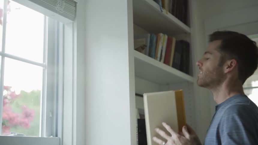 A man takes a book down from a shelf near a window