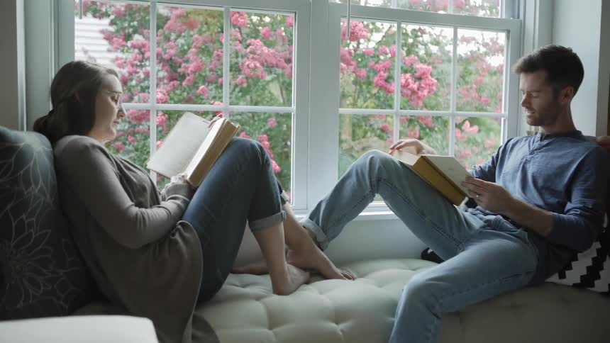The two people sit by the window in their living room reading