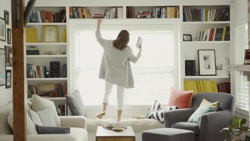 Woman stands on a sofa and reaches up into the bookshelf above