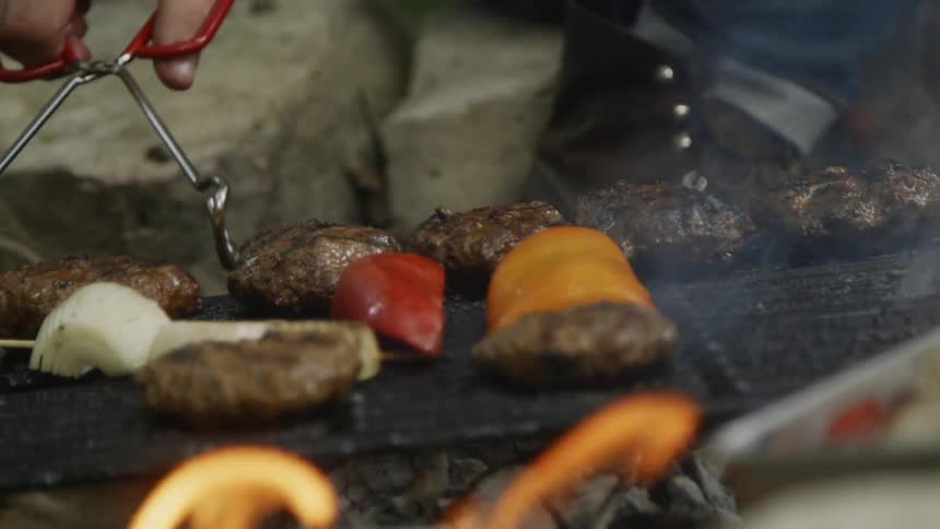 A person uses tongs to adjust burgers cooking on a flaming grill