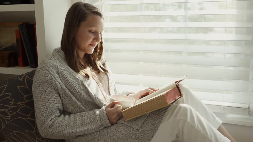 Woman sits on a couch next to a window reading a book