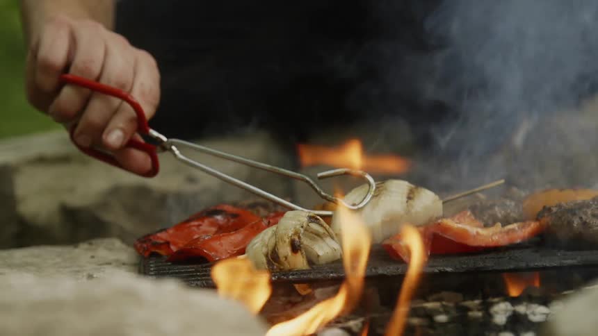 A person uses tongs to flip onions on a flaming grill