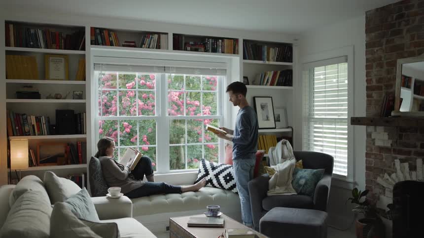 Man gets a book from the shelf as a woman sits on the sofa reading