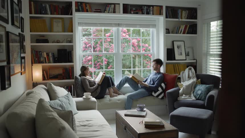 Man and woman sit reading on a couch next to a large window