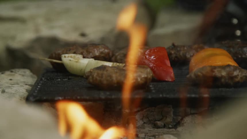 Peppers onions and burgers cooking on a grill with flames outside