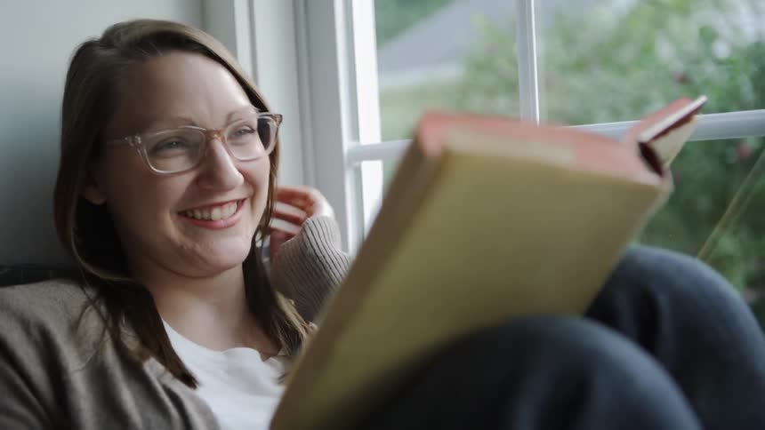 Woman laughs as she sits next to a window reading a book
