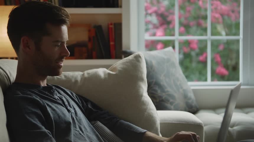Man smiles as he sits on a couch typing on a laptop