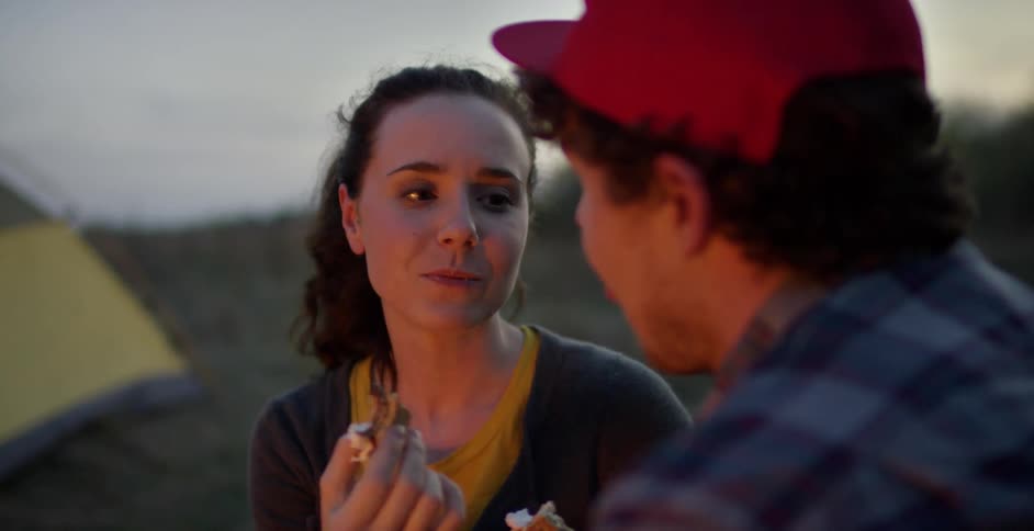 A man and woman talking and eating by a campfire