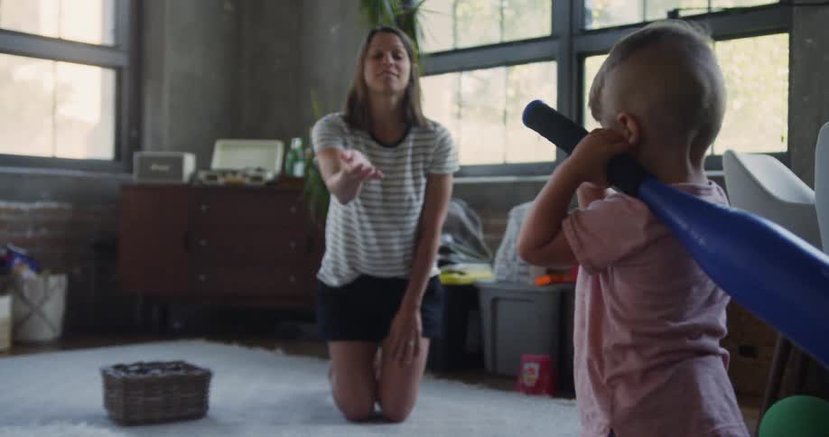 A woman kneels on the floor and throws a ball to a little boy who swings a plastic bat and tries to hit it