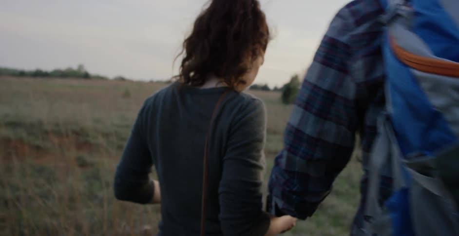 A man wearing a backpack holding the hand of a brunette woman as they walk through a field