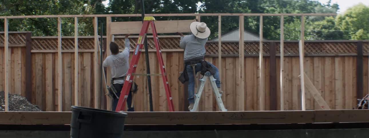 Two men work to put up a piece of a wood on the frame of a building near some trees and a fence
