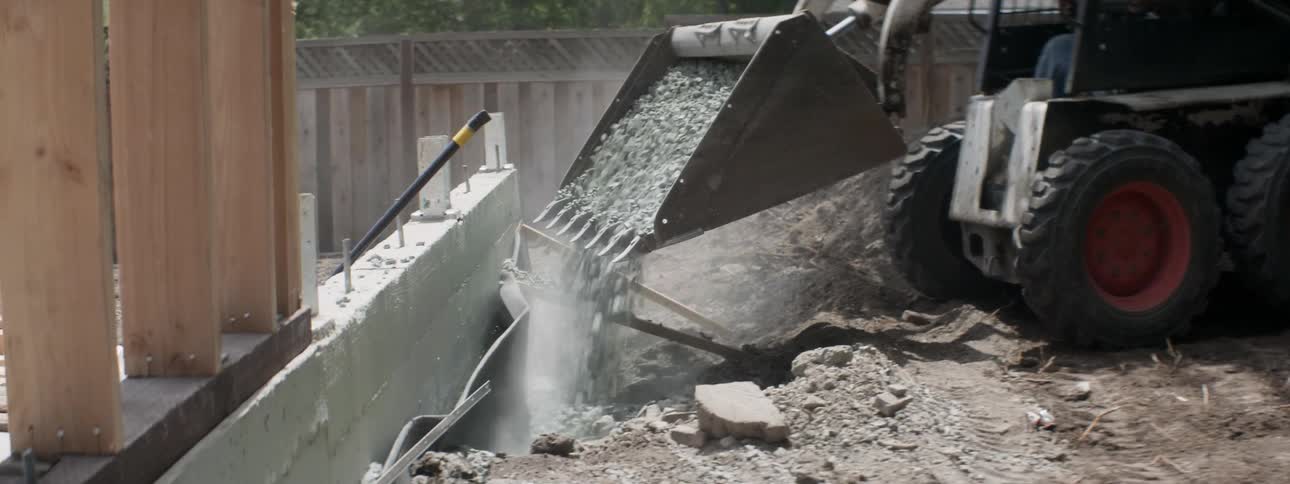 A front loader drops gravel near a new cement foundation wall
