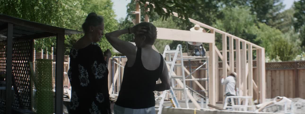 Two women stand and talk while two men work on a building