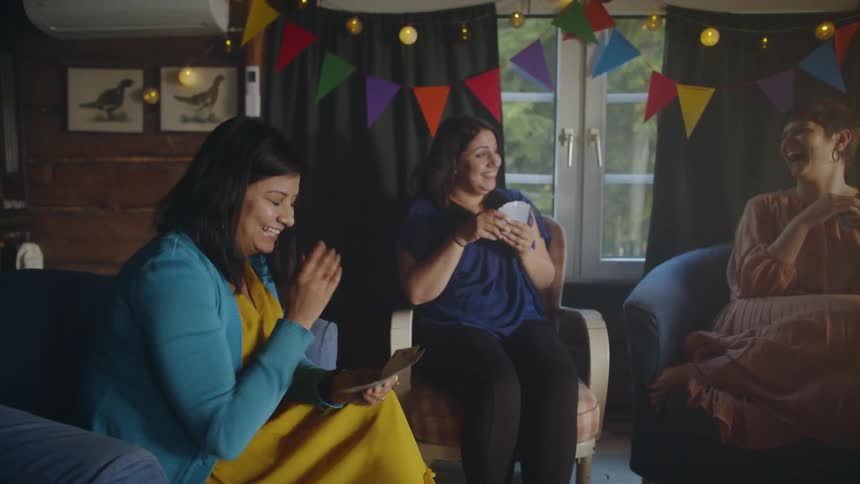 A group of women sit in a room decorated for a party laughing and playing cards