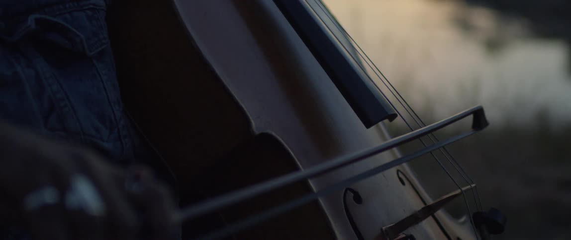 A woman stands outside by a body of water at sunset playing a cello