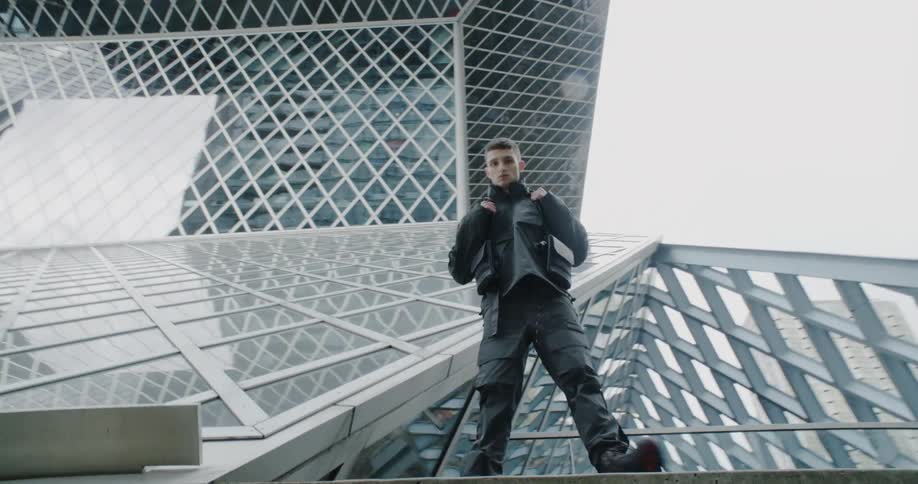 A man wearing a vest with pockets stands and poses on a large building on a sunny day