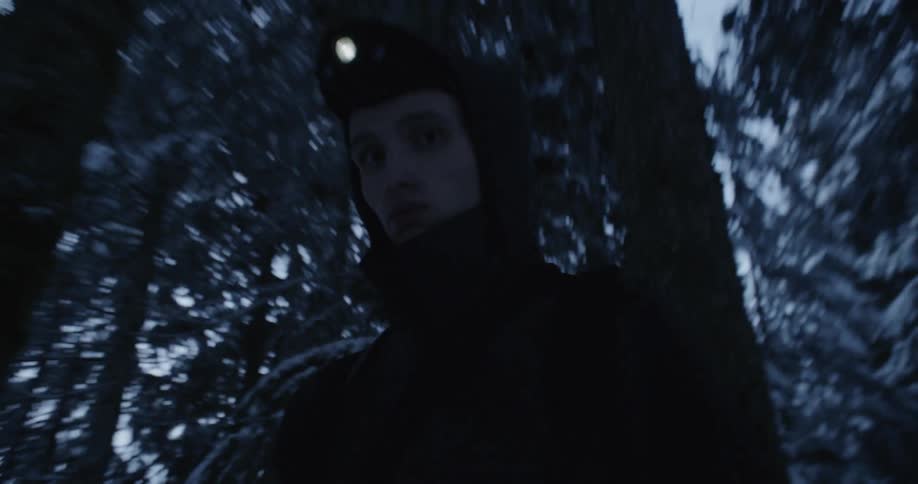 A man wearing a headlamp walks through a forest at dusk