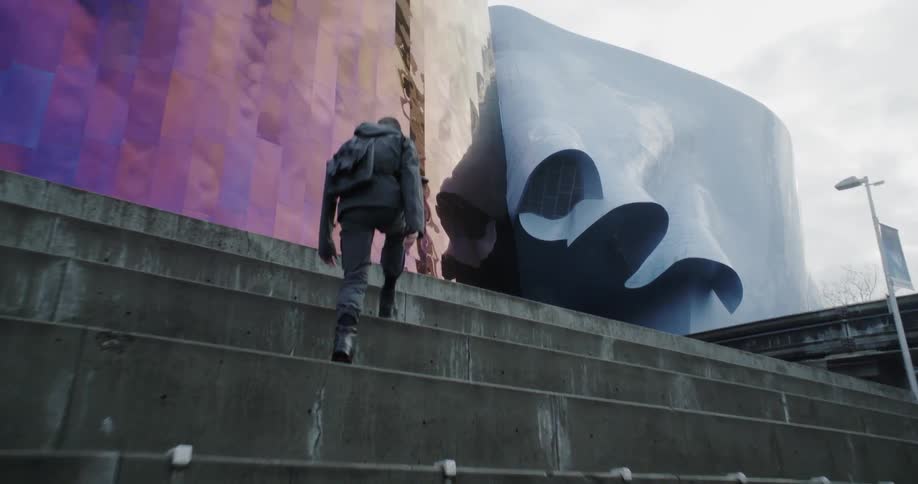 A man climbs up the steps to a large iridescent building on a cloudy day