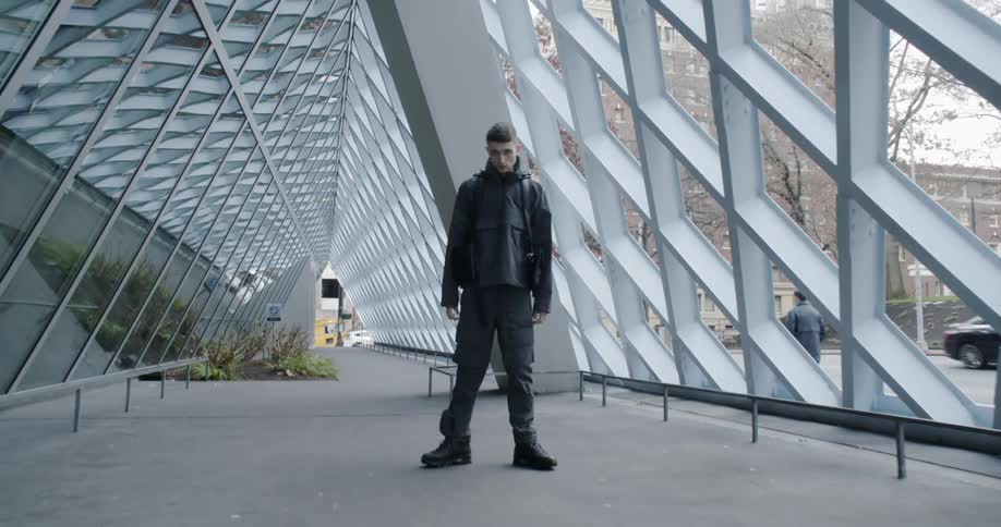 A man stands in front of a large building as cars drive by