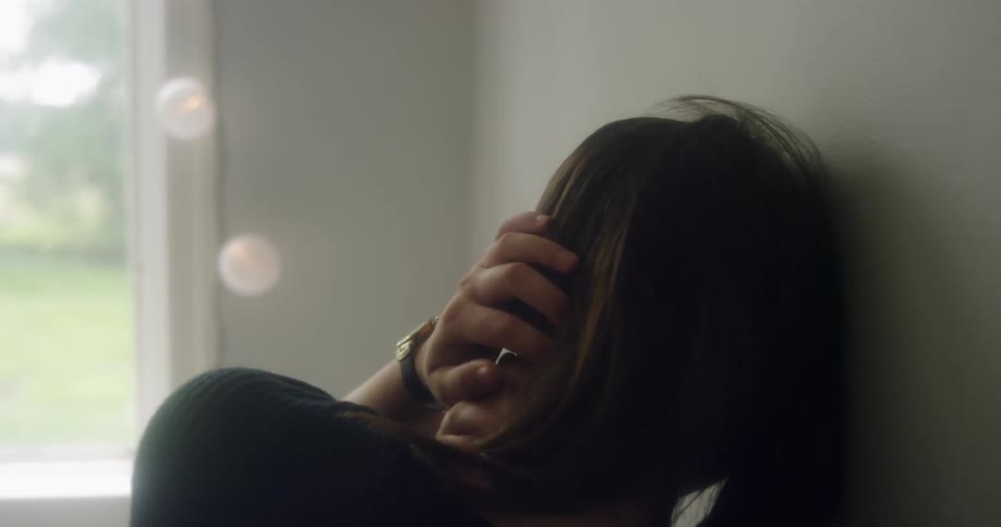 A woman leans against a wall near a window and strokes her hair
